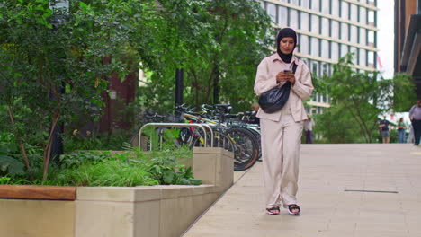 Muslim-Businesswoman-Wearing-Hijab-Going-To-Work-Standing-Outside-Modern-Office-Looking-At-Mobile-Phone-2