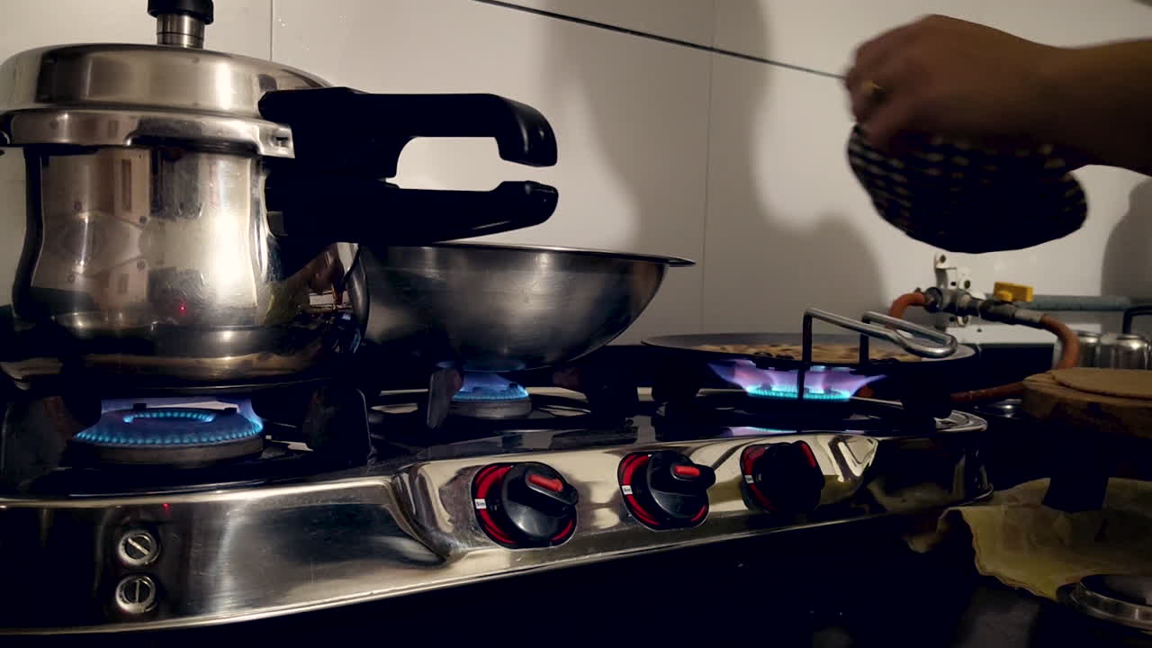 Person cooking on gas stove with blue flame, steel utensils and pressure cooker in home kitchen. Useful for household, food preparation and daily lifestyle content