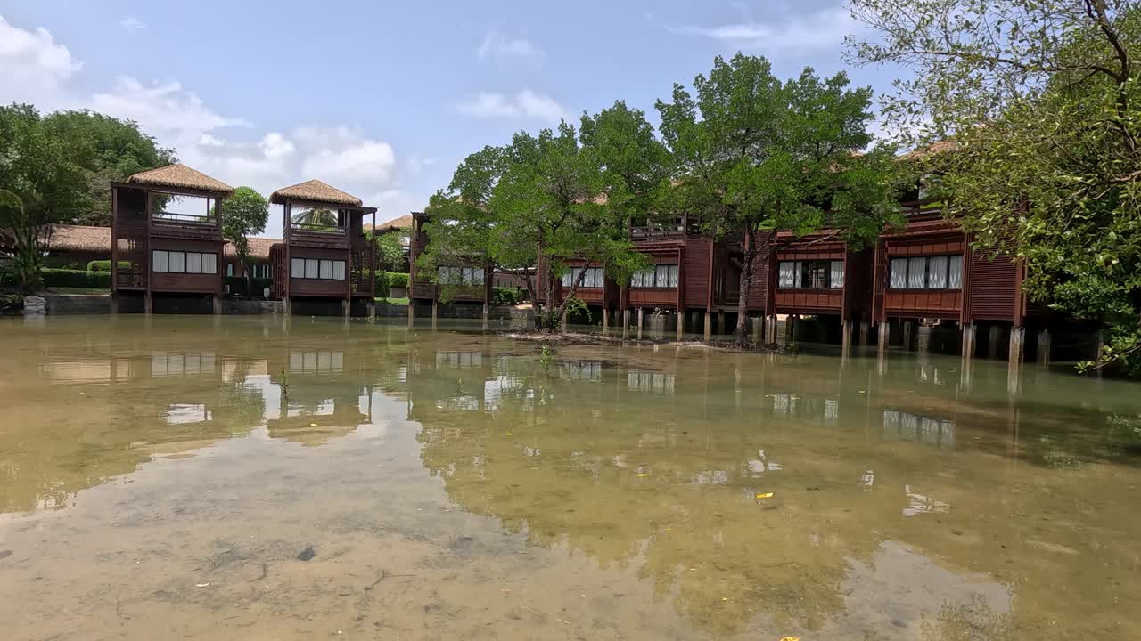 Camera moves steadily from a thatched overwater bungalow through a mangrove wetland, revealing wooden villas, lush greenery, and a sandy beach under bright daylight