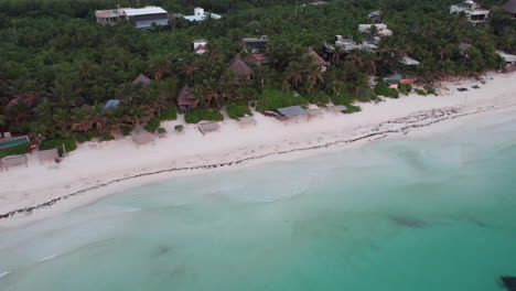 Aerial-Tilt-down-shot-of-a-white-sand-beach-with-crystal-clear-blue-ocean-with-huts-and-cabins-surrounded-by-palm-trees-in-Tulum,-Mexico