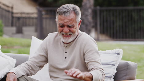 Senior-Hispanic-man-sitting-on-a-sofa-in-the-garden-laughing-to-camera,-waist-up,-close-up