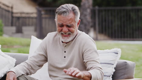 senior hispanic man sitting on a sofa in the garden laughing to camera, waist up, close up