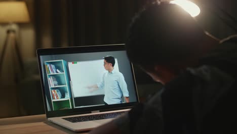 school teen boy student distance learning with online teacher on computer screen. web tutor gives remote class teaching teenage pupil elearning from home at night. over shoulder close up view