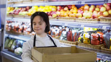 retrato de una niña con síndrome de down que sostiene una caja con mercancías con estantería de frutas en el fondo