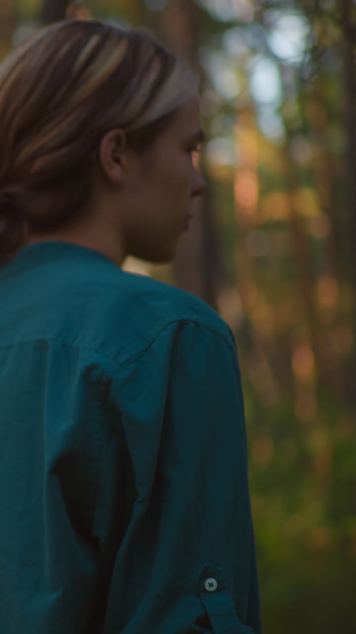 mujer joven pensativa en camisa verde, llevando una bolsa roja, camina a través del bosque iluminado por el sol, bañado en un brillo cálido de la luz del sol filtrando a través de árboles altos en el fondo
