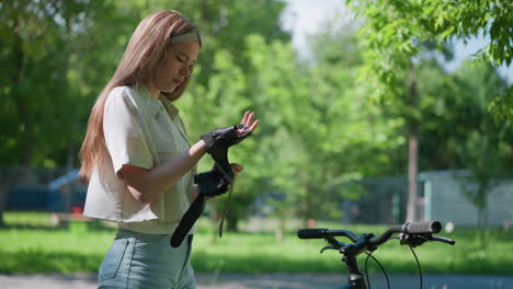 young woman standing outdoors near bicycle, fastening biker glove on her right hand with a focused expression, background features blurred trees, lush greenery, and a fence