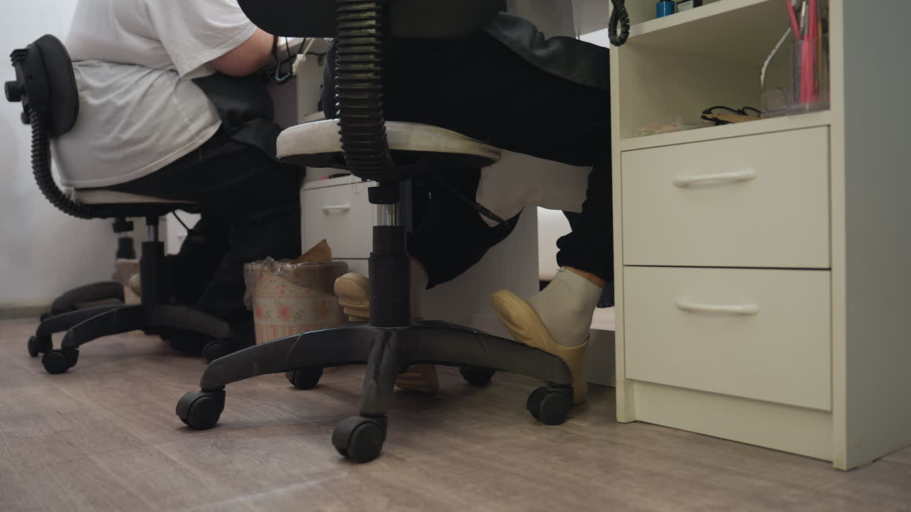 Salon interior floor-level view featuring black swivel chair with wheels and technician wearing beige slippers, with nearby workstation shelves, cosmetic supplies, and floral-patterned trash bin