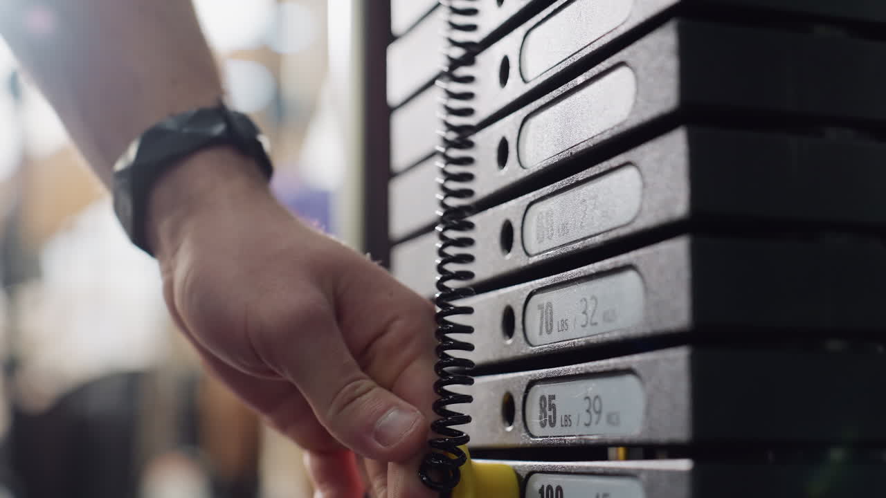 Student hand inserts pin into lower weight plate on cable machine stack in modern gym, precise adjustment ensuring correct resistance before workout, showing preparation and safety in gym routine