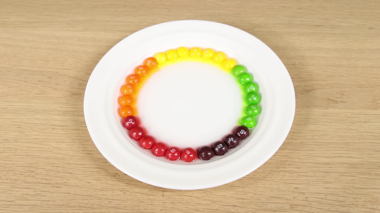 Candies dissolve in water, creating a vibrant radial pattern on a white plate under bright lighting
