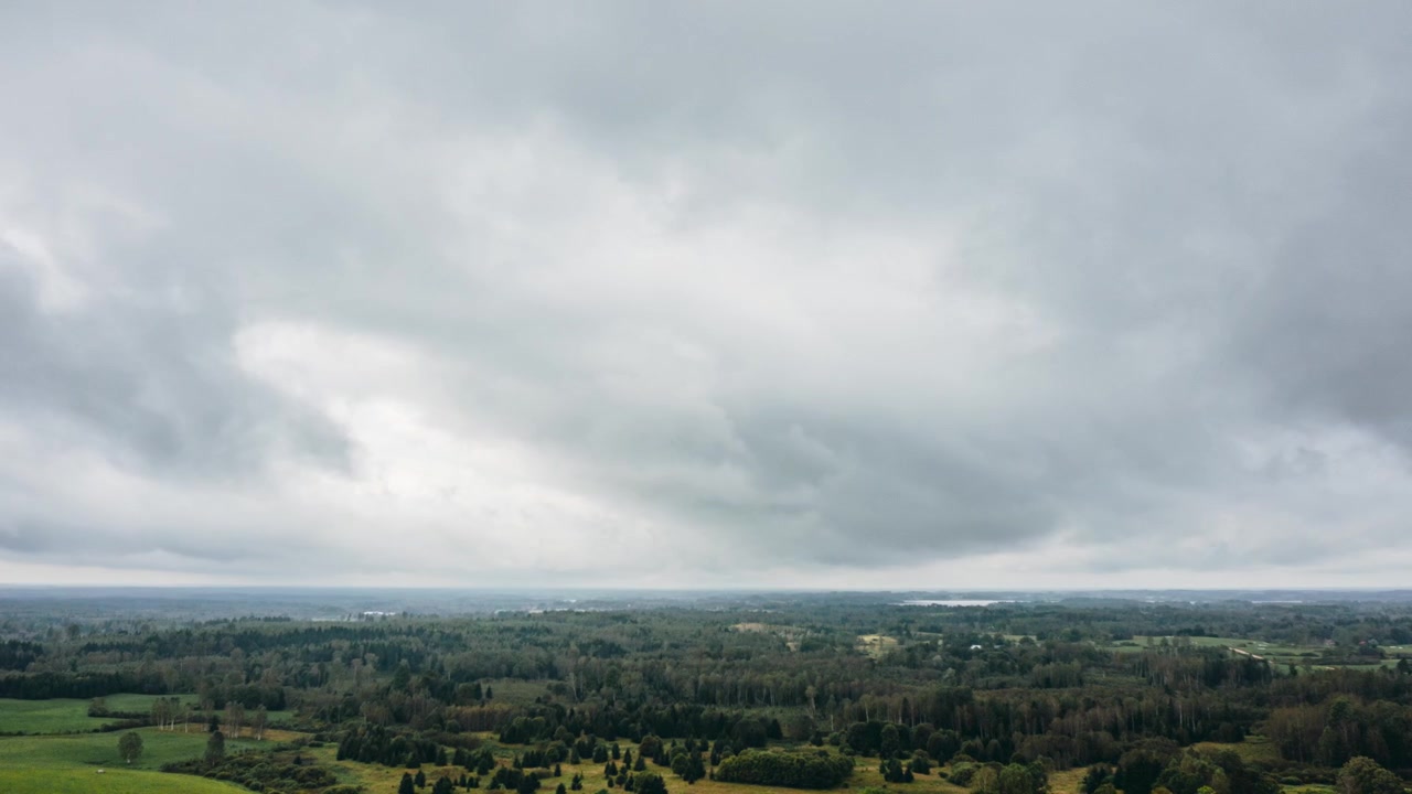 Hyperlapse dronevisning fra luften av tunge grå stormskyer som beveger seg over et vidstrakt grønt landskap med skog og bølgende jorder.