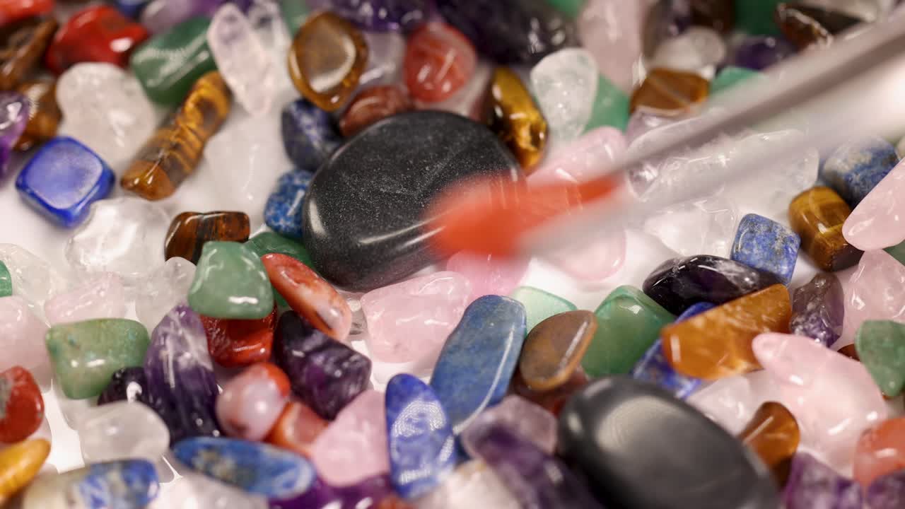 Tweezers carefully pick a red jasper stone from a vibrant assortment of chakra stones. Close-up view with focused lighting