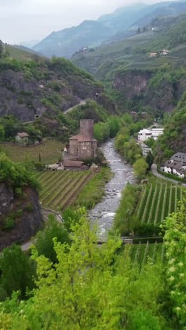 vertical drone shot flying toward roncolo castle above green vineyards and a small river
