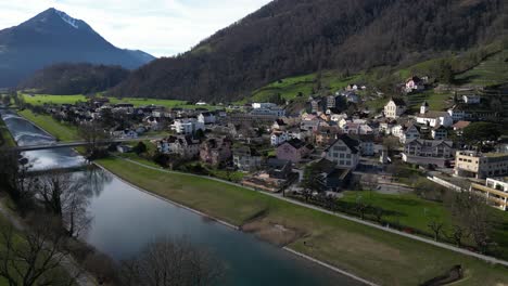 Panning-drone-clip-of-scenic-Swiss-village,-with-snowcapped-mountains-in-the-background-and-calmly-flowing-river-with-bridge