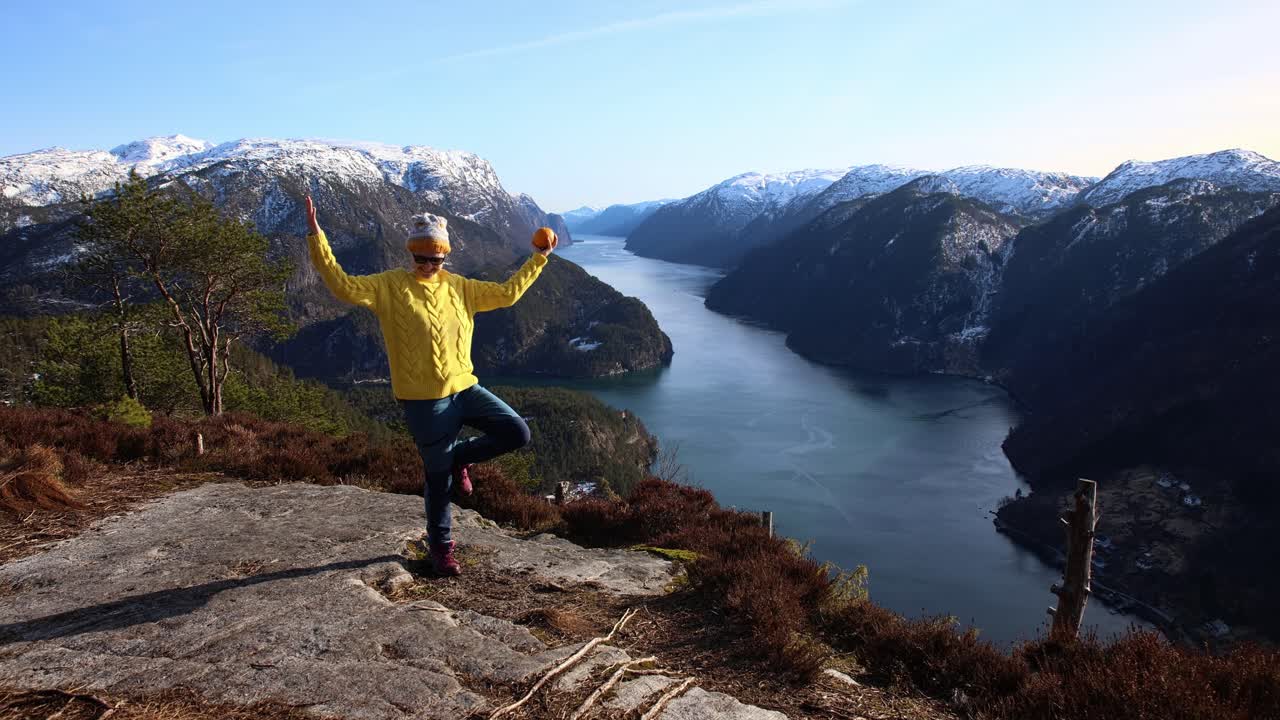 feliz chica noruega en suéter de pascua amarillo sosteniendo una naranja en la cima de la montaña con impresionante vista del fiordo - veafjord noruega