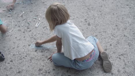 back view of blonde girl sitting on asphalt and drawing with chalks