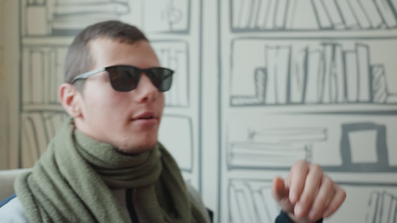 White man in sunglasses on sofa, cozy library wallpaper backdrop, green scarf, flipping notebook, checking watch, thoughtful expressions, indoor natural light, warm bokeh, candid closeups