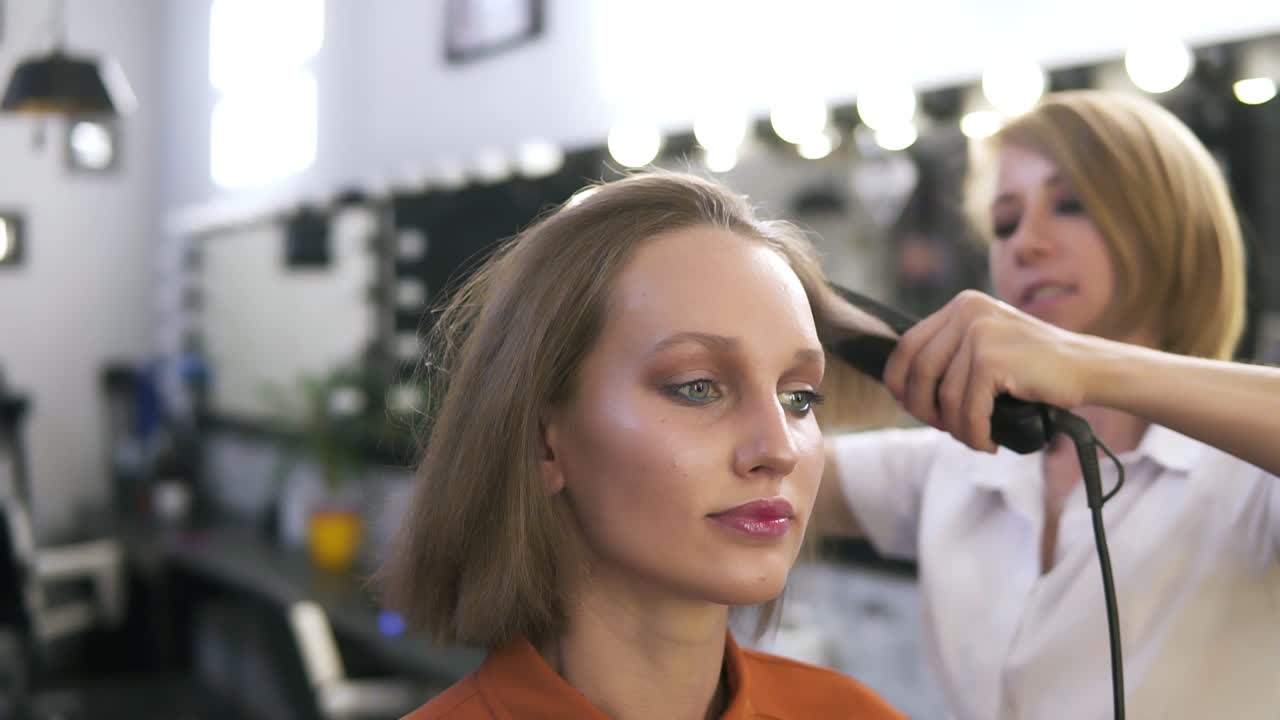 Attractive caucasian girl with make up in beauty salon. Hairdresser straightens her hair with and iron. Blurred background -