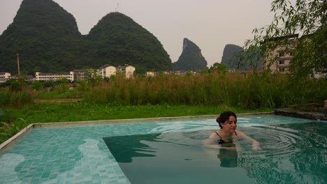 Caucasian woman swimming in pool with karst mountains and thin cloudy sky in background. Yangshuo, China