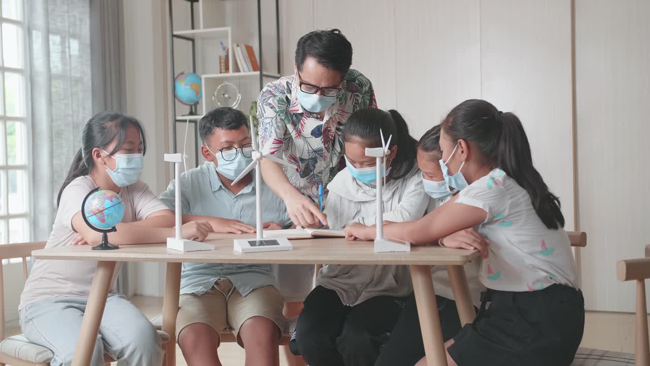 Middle School Classroom, Wearing Facemasks , Enthusiastic Asian Teacher Explains To Young Children How Wind Turbines Work. Kids Learning About Eco-Friendly Forms Of Renewable Energy