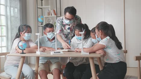 middle school classroom, wearing facemasks , enthusiastic asian teacher explains to young children how wind turbines work. kids learning about eco-friendly forms of renewable energy