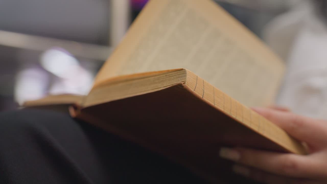 Close up of person holding open book and gently flipping page with fingers, wearing white shirt and sitting comfortably indoors with soft blurred background
