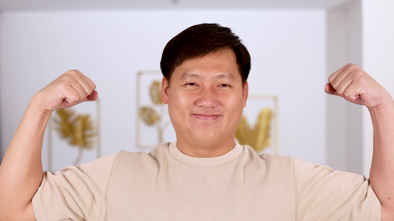 Asian man in beige shirt flexes arms and changes facial expressions in a bright, modern indoor setting with soft, even lighting and steady camera