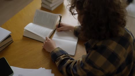 over the shoulder a girl with curly hair in a plaid shirt takes notes on a book while reading in the library