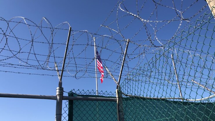 Bandiera a stelle e strisce degli Stati Uniti contro il cielo blu come striscione per una struttura americana recintata con grata, filo spinato e barriere di filo spinato