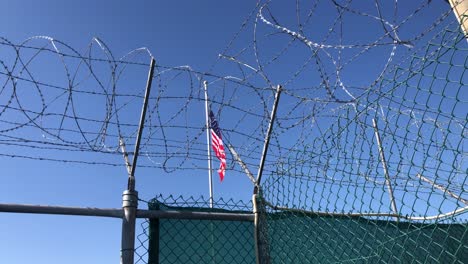 The-United-States-stars-and-stripes-banner-against-blue-sky-as-banner-for-a-fenced-american-facility-with-grate,-razor-wire-and-barb-wire-barriers