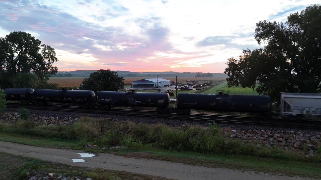 A drone tracks alongside a long freight train hauling tanker and hopper cars as it rolls through open farmland beneath a fading pink sunset sky near a quiet country road.
