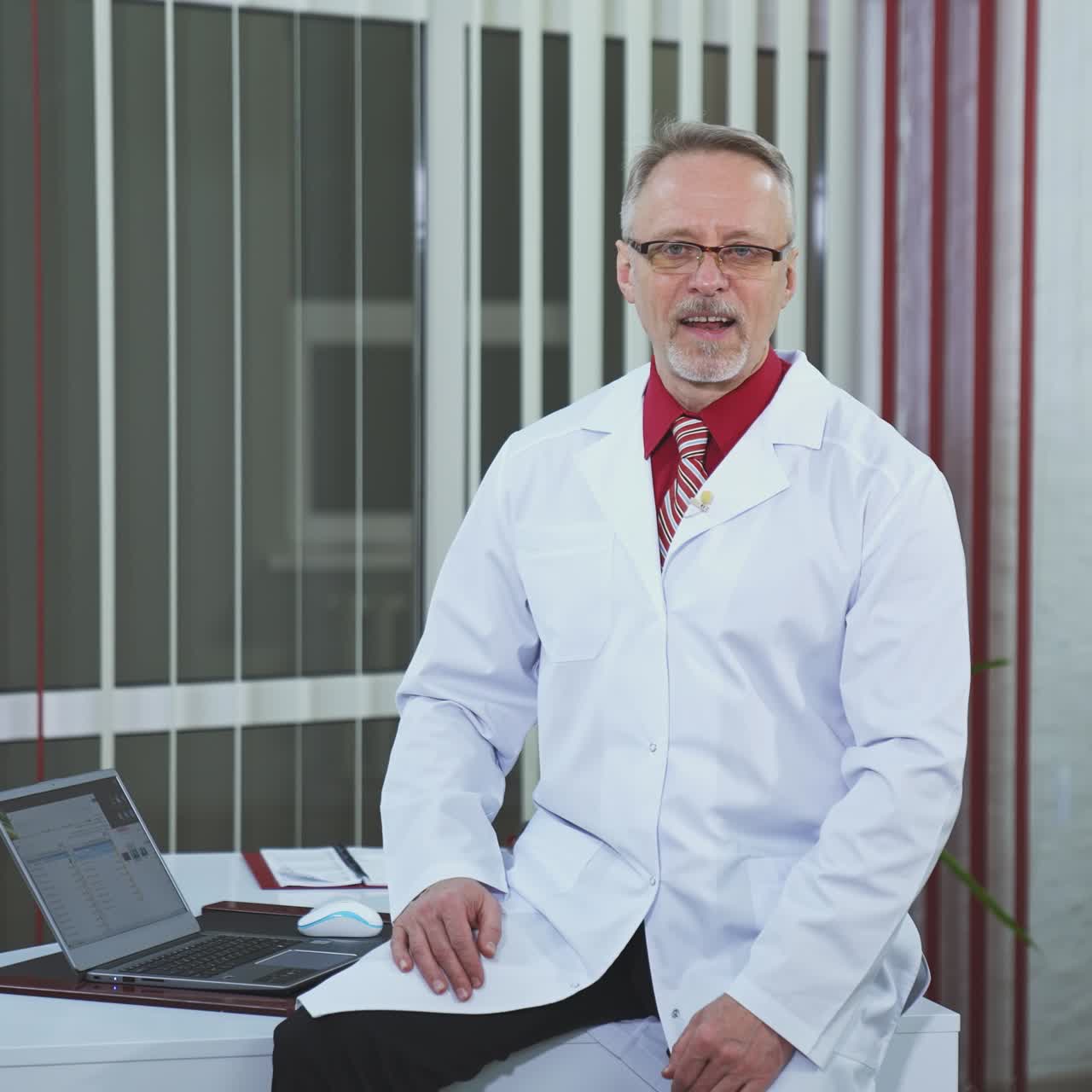 Physician recording webinar training. Professional male doctor sitting on desk and talking in healthcare center. Doctor in glasses and white medical uniform looking at camera.