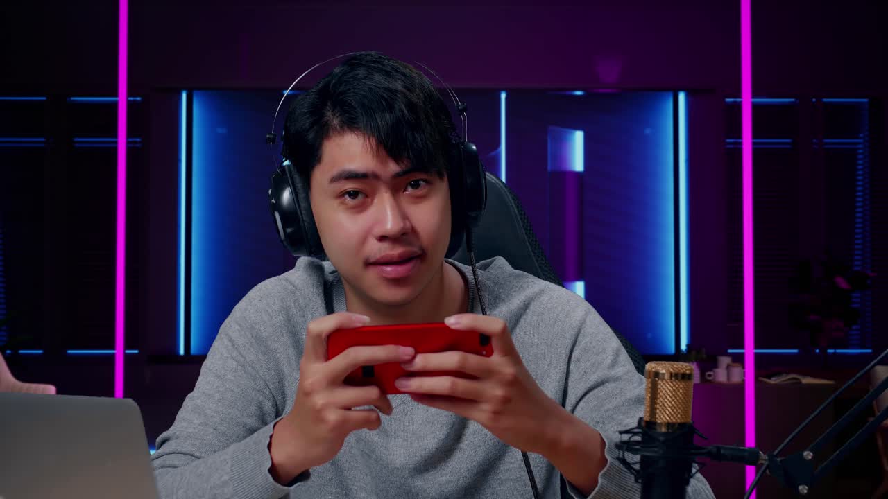 헤드폰과 컴퓨터를 가진 미소 짓는 아시아 남성 게이머가 레트로 네온 조명으로 조명된 방에서 휴대 전화 게임을 하는 동안 카메라를 바라보고 있습니다.