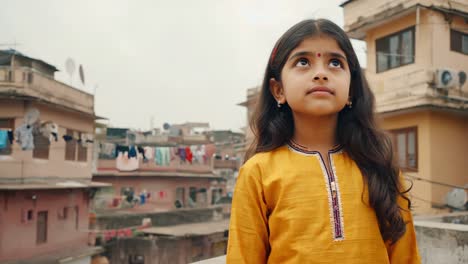 young indian girl in traditional clothing looking up with a thoughtful expression, standing on a rooftop with clothes hanging to dry in the background, symbolizing hope and dreams for the future