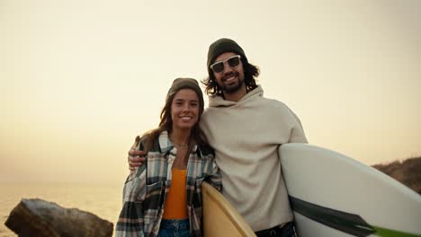 Portrait-of-a-happy-couple,-a-brunette-man-in-a-white-sweatshirt-and-black-sunglasses-together-with-his-blonde-girlfriend-in-a-plaid-shirt-stand-near-the-sea-and-hold-surfboards-in-their-hands-in-the-morning-in-autumn