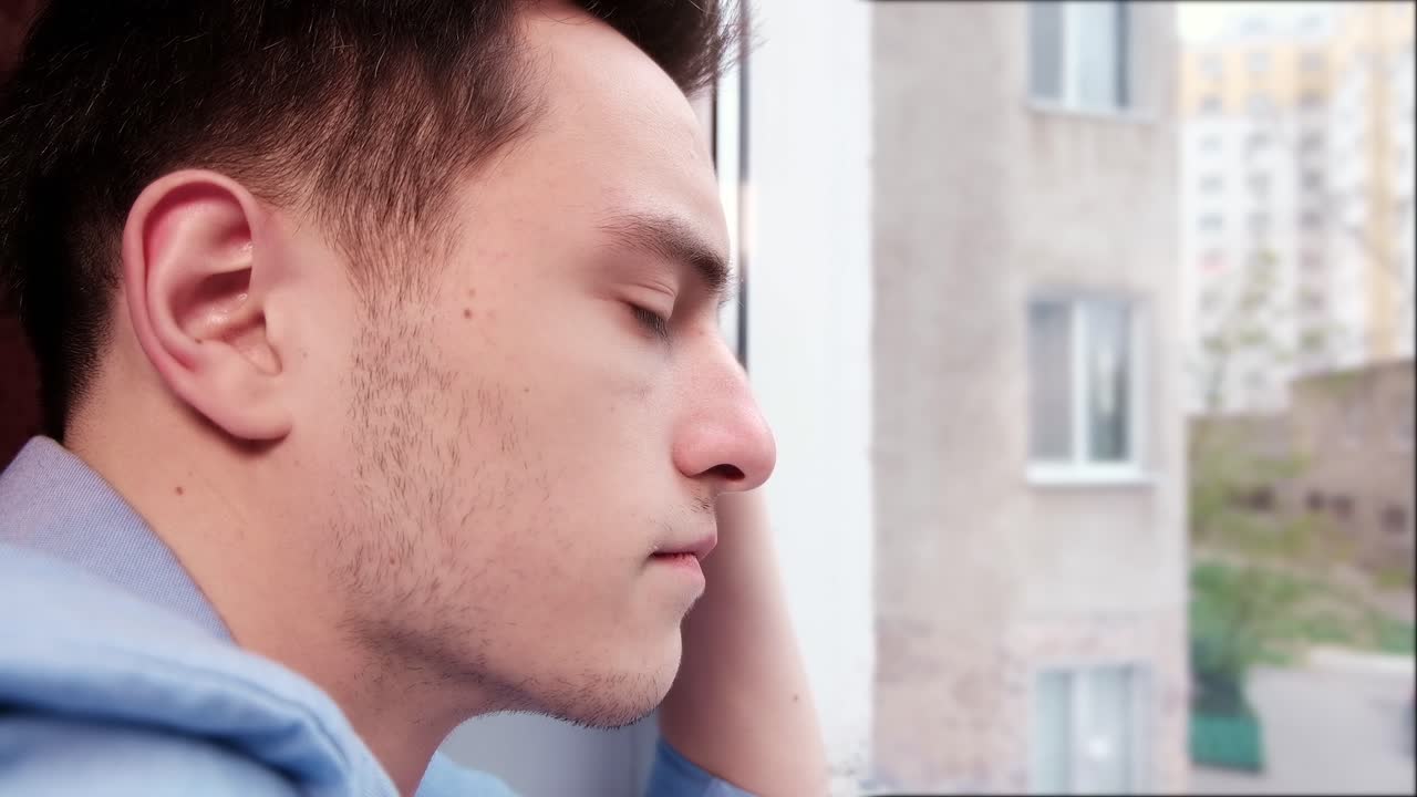 Bored man in blue shirt looking desperate out of the window while keeping hand on the cheek. Close up. Quarantine and Corona Virus idea