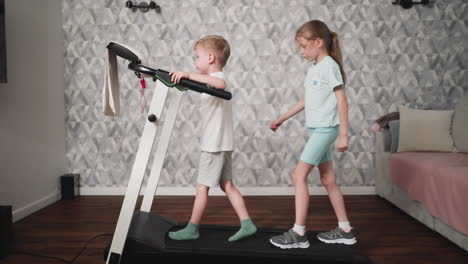 girl focuses on maintaining balance while walking on treadmill behind boy who looks downward with serious expression, both children involved in exercise session inside cozy room with soft furniture