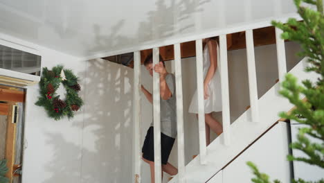 kids coming down white staircase, smiling and very happy, boy in gray top and black shorts, girl in white gown, festive indoor setting with holiday decorations, family moment, fun and excitement