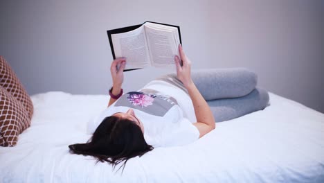 a young woman is reading a book with black hardcover in her bedroom, holding boom in the air, laying on the bed, she spends her free time with a book, relaxing at home, turns the page, flips the page