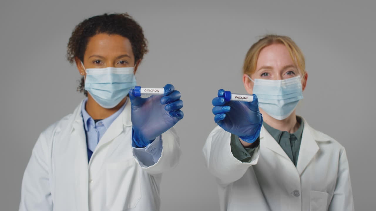 Studio Shot Of Lab Research Workers In Face Masks Holding Test Tubes Labelled Vaccine And Omicron