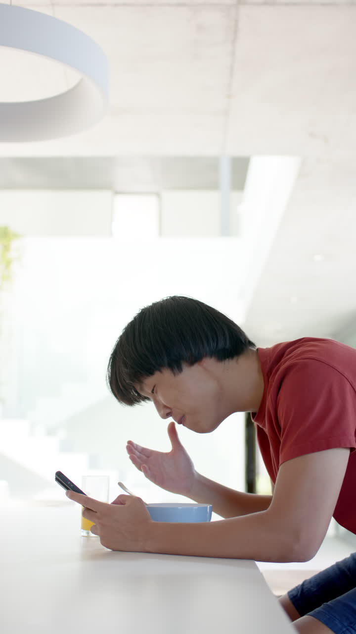 Vertical video: Asian teenage boy eating breakfast and using smartphone at home, looking surprised