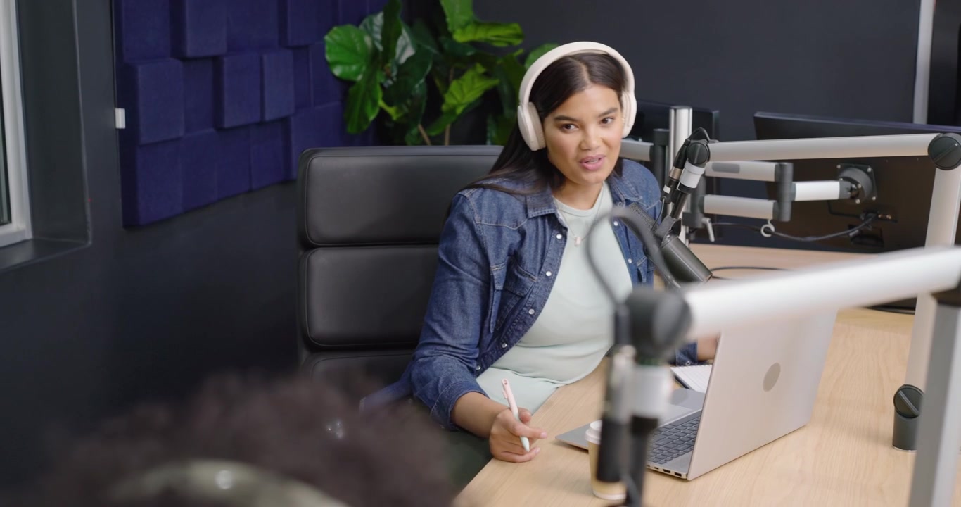 Co-apresentadoras diversas, uma de camisa rosa ajustando o microfone, apresentadora sentada de jeans falando no laptop