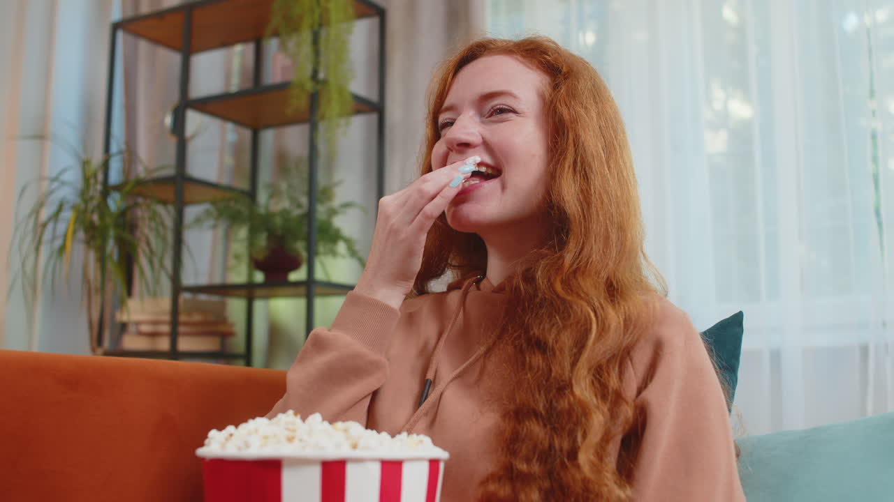 улыбающаяся рыжая женщина ест попкорн и смотрит фильм wow выражение лица сидит на диване дома