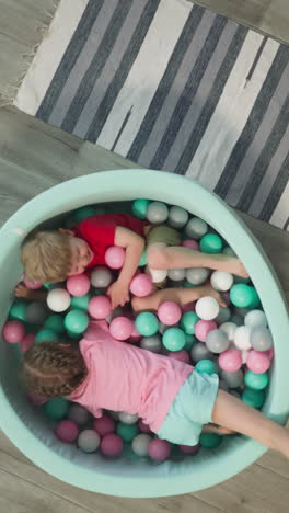 glad couple of children wearing t-shirts and shorts crawls in dry pool. little sister and brother move under plastic balls smiling joyfully