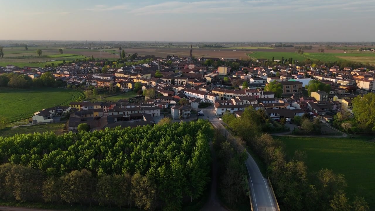 Villaggio di Spinadesco vicino a Cremona, Lombardia, Italia, con edifici residenziali, torre campanaria e i campi agricoli intensivi circostanti nella Pianura Padana, ripresa aerea con drone in ascesa che svela il panorama.