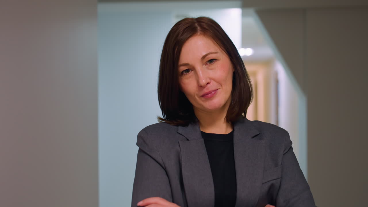 Confident professional woman in formal blazer standing with arms crossed and holding notebook in modern office corridor, expressing authority, calmness, and readiness for business or leadership tasks