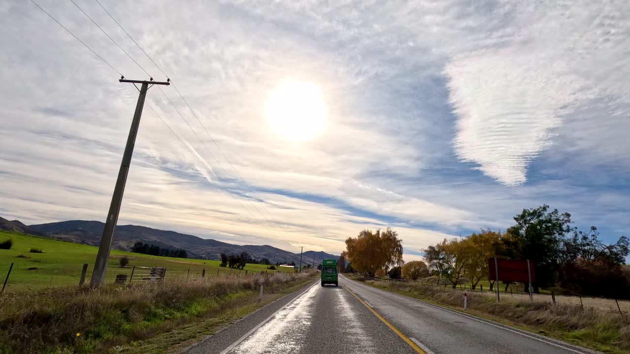A campervan travels a sunlit road in Queenstown, New Zealand, surrounded by autumn foliage and expansive landscapes