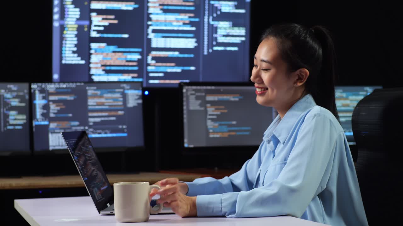 vista lateral de una programadora asiática bebiendo café mientras escribe código en una computadora portátil usando múltiples monitores que muestran la base de datos en los escritorios de ventana de terminal en la oficina