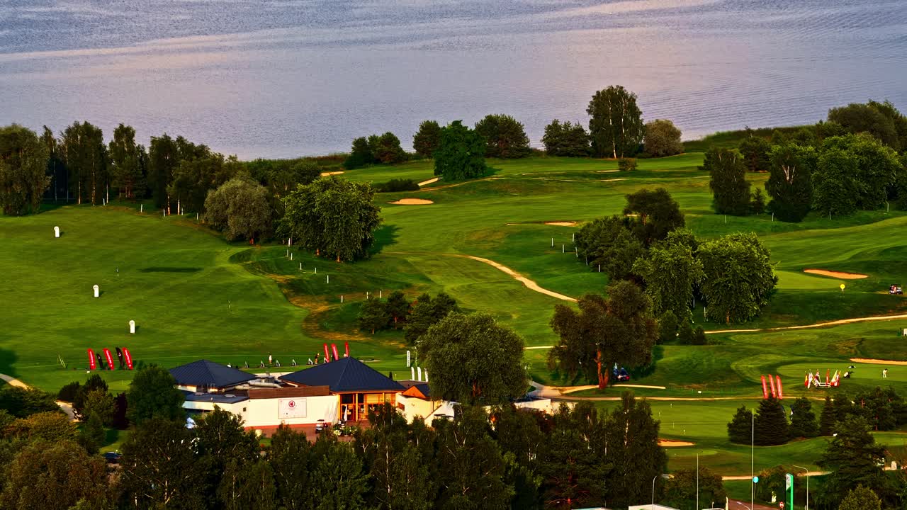 Aerial view of lush green golf course alongside a lakeshore, with fairways, bunkers, and surrounding trees under golden sunset light