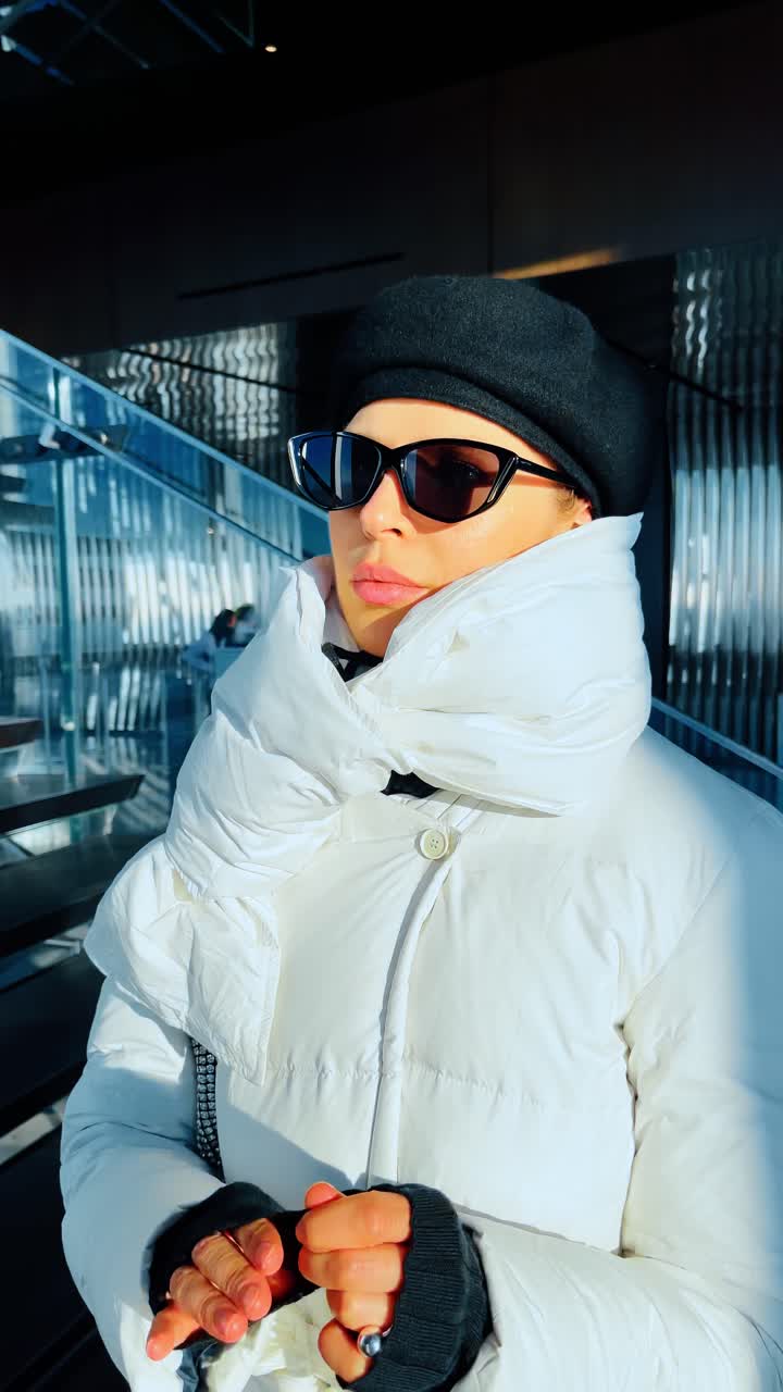 New York, USA, 25 December 2022: Woman in white winter coat inside modern tower. Woman in white puffer coat, black beret, sunglasses, glass stairs, direct sunlight, New York interior
