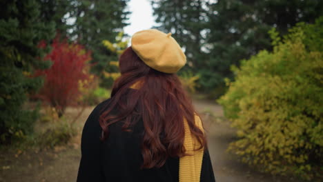back view of artist with long wavy red hair wearing a black coat, yellow beret, and matching scarf, adjusting her hair while walking through a lush green park adorned with colorful autumn foliage